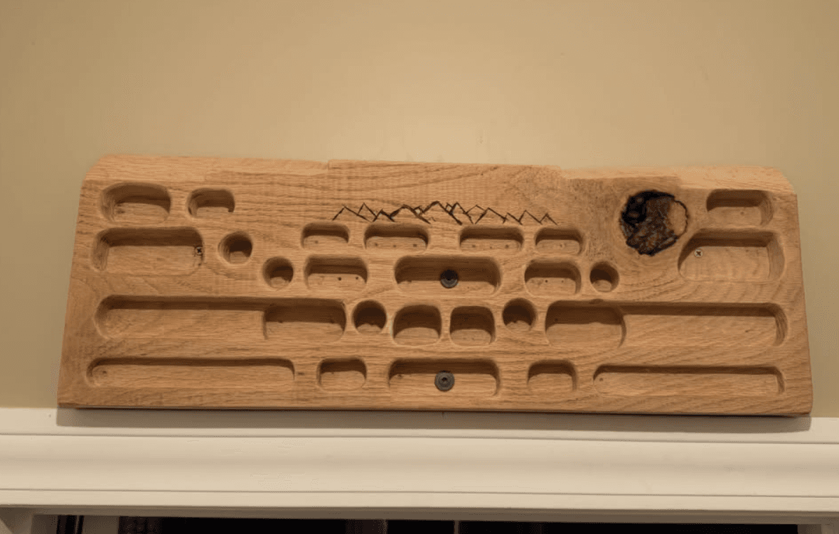 Hand-routed oak hangboard mounted on a wall, showing four rows of edges and pockets with a woodburned mountain range across the face