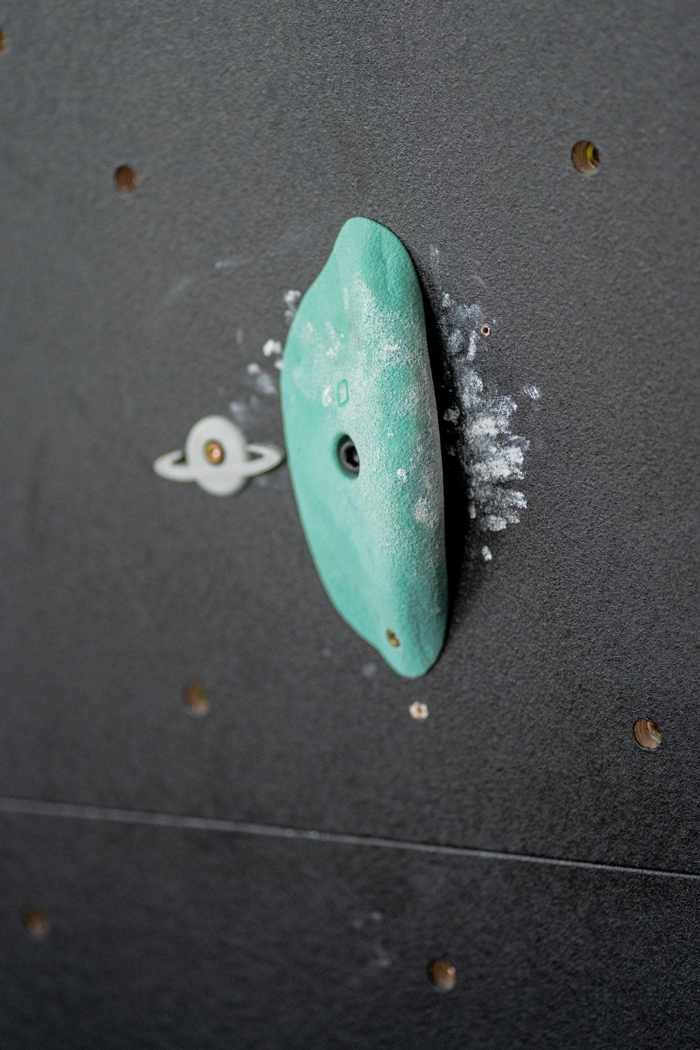 A ringed-planet route tag mounted beside a chalked green climbing hold on the gym wall