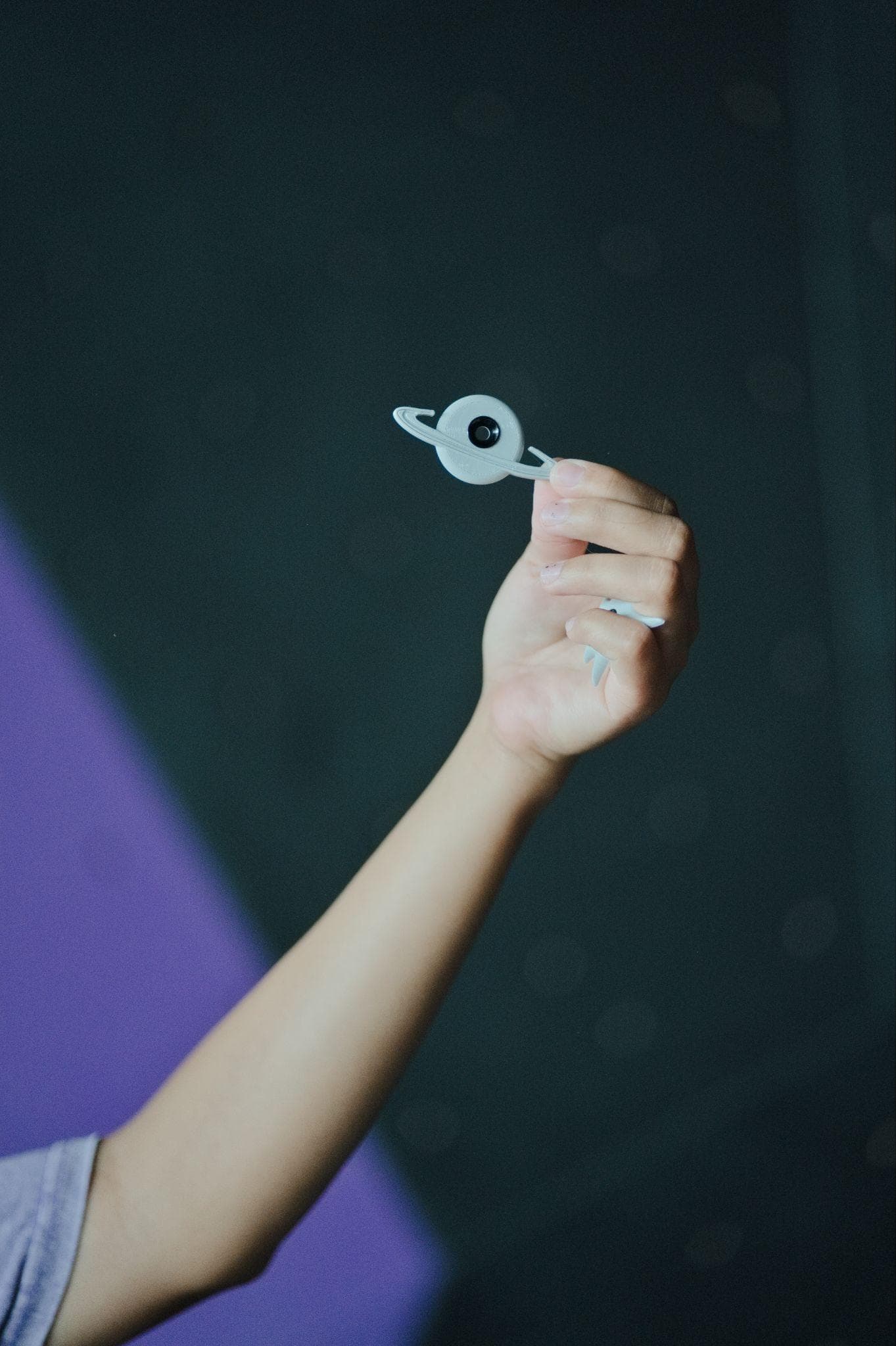 A hand holding up a ringed-planet route tag in front of a dark climbing wall