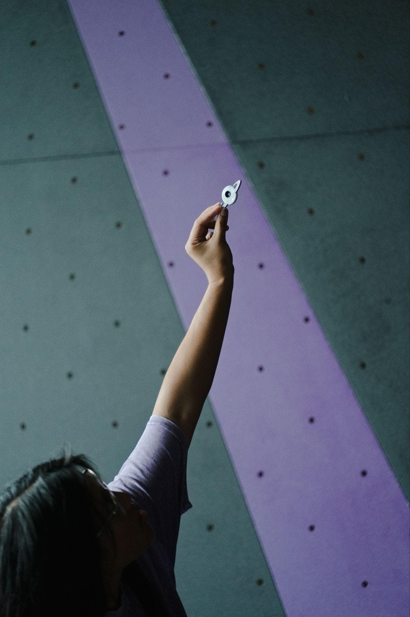 A climber reaching up with a route tag toward a purple and teal bouldering wall